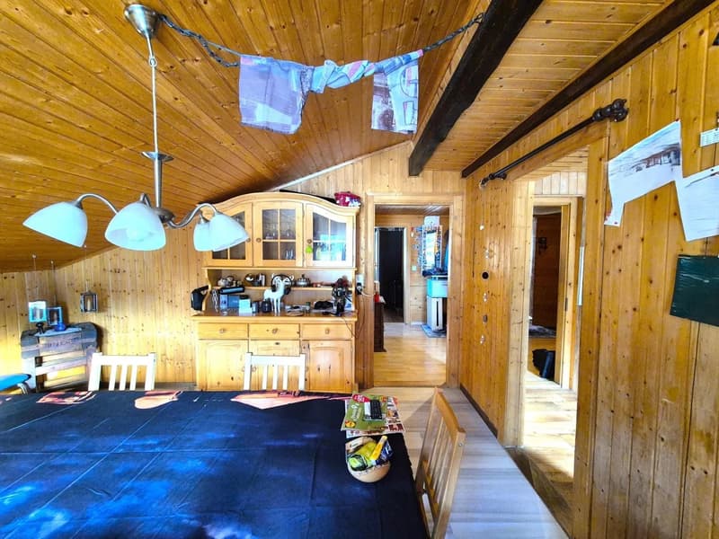 The image shows a cozy, rustic interior with wooden walls and ceiling. There is a kitchen area with a wooden cabinet and shelves, as well as a dining table and chairs. Laundry items are hanging from a line, indicating the presence of a washing machine. Th