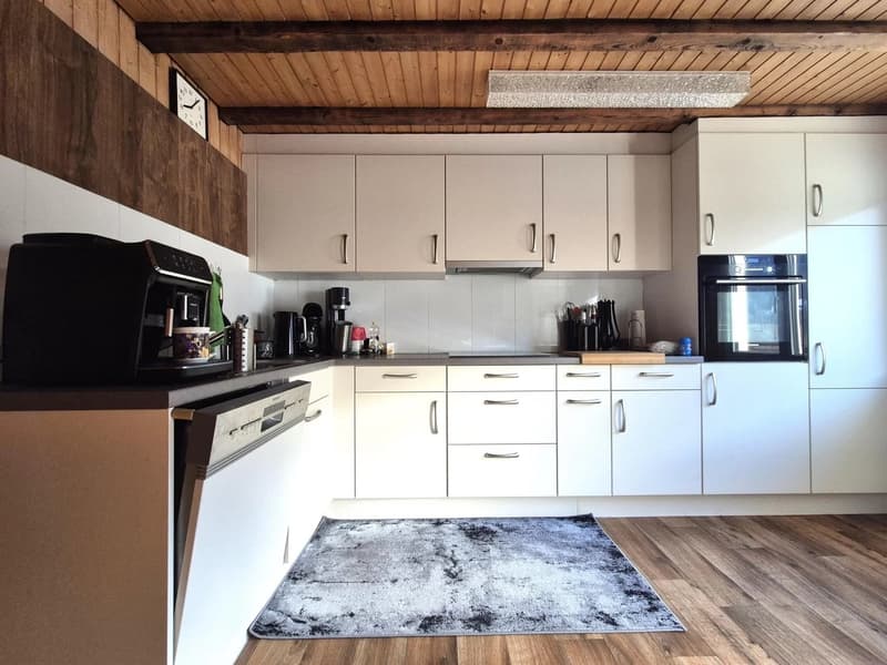 The image shows a modern kitchen with white cabinets, a black oven, and various kitchen appliances such as a coffee maker and a refrigerator. The kitchen has a wooden ceiling with exposed beams and a wooden accent wall. The floor is made of hardwood, and
