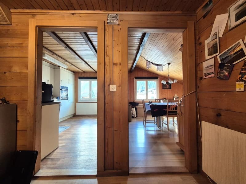Rustic wooden cabin interior with open floor plan, exposed wooden beams and paneling, large windows, dining area with table and chairs, and a fireplace visible in the background.