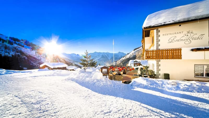 Snow-covered mountain landscape with a traditional Alpine-style restaurant building, outdoor seating area, and snowy terrain in the foreground