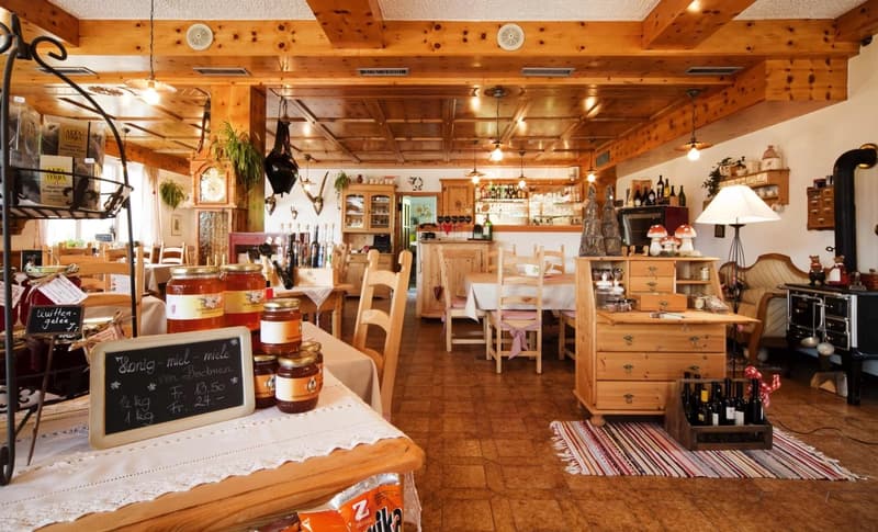 A restaurant interior with wooden ceiling, wooden floor, wooden tables, wooden chairs, white tablecloths, jars of honey, blackboard with text, wooden wine cabinet, white lamps, shelves with bottles, bottles, a gas stove, a plant on the wall, and a cabinet