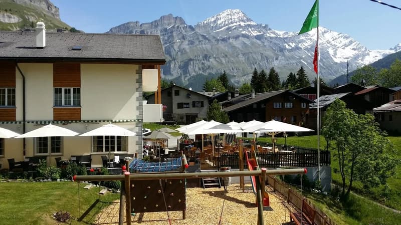 The image shows an outdoor seating area of a restaurant or cafe in a picturesque mountain setting. There are white umbrellas covering the tables, and the building in the background appears to be a hotel or resort. The snow-capped mountains in the distance