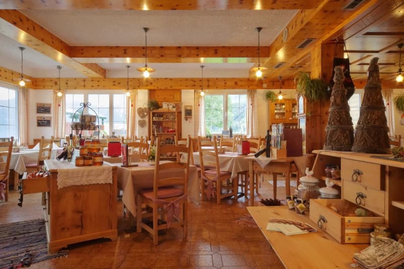This image shows a cozy, rustic-style restaurant or cafe interior. The space features wooden beams and paneling, giving it a warm and inviting atmosphere. There are several dining tables with chairs, as well as shelves and decor items along the walls. A f