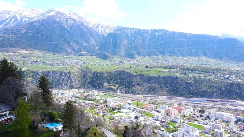 The image shows a panoramic view of a mountainous landscape. In the foreground, there is a town or city nestled in the valley, surrounded by lush green forests and snow-capped peaks in the background. The scene appears to be a picturesque alpine setting.