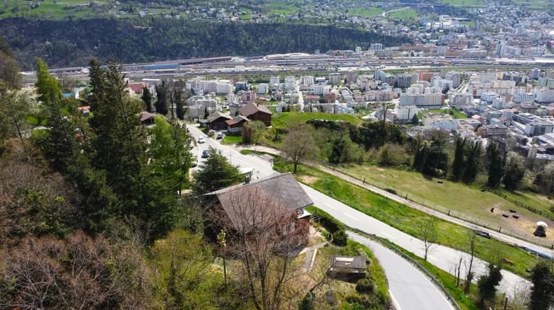 Aerial view of a mountainous city with a mix of residential and commercial buildings, surrounded by lush greenery and forests. The city appears to be situated near a railway line or transportation hub. The landscape features a winding road leading through