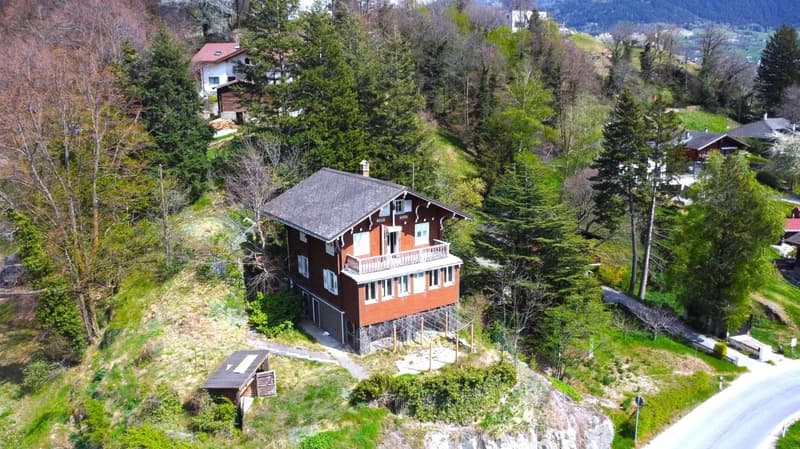 2-story chalet-style house with a red roof, surrounded by lush greenery and trees, situated on a hillside with a view of the mountains and valley below