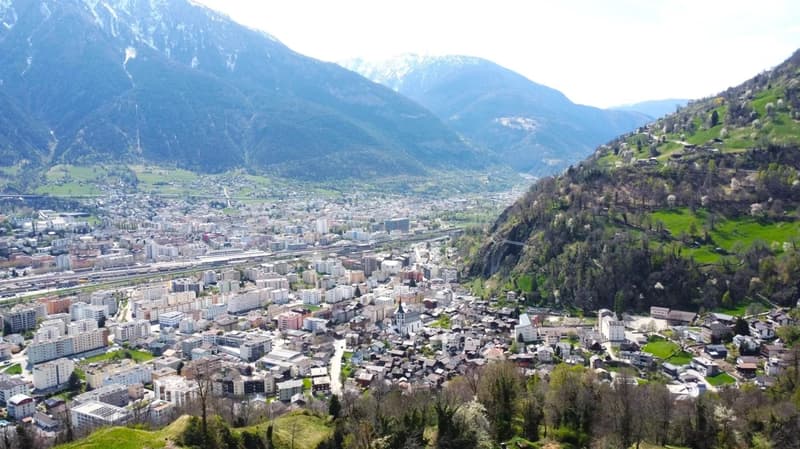 Aerial view of a mountainous city with a mix of high-rise buildings, residential areas, and green spaces. The city is surrounded by towering mountains and lush vegetation. There appears to be a river or stream running through the center of the city.