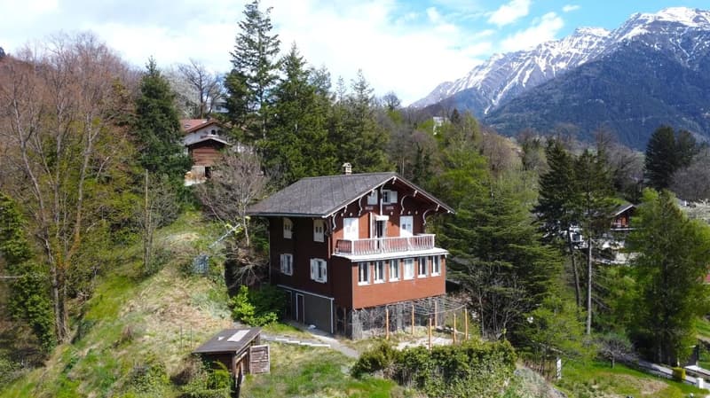 2-story wooden chalet-style house with a balcony, surrounded by pine trees and mountains in the background