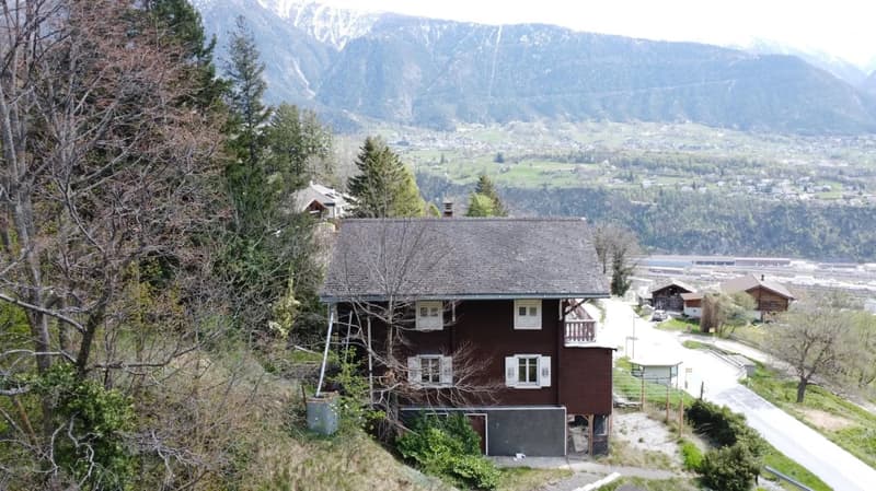 A traditional wooden chalet-style house nestled in a mountainous, forested landscape with a view of a town or village in the valley below.