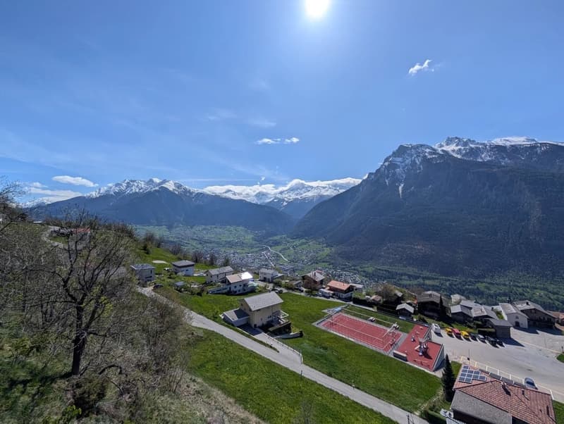 The image shows a scenic mountain landscape with snow-capped peaks in the background. In the foreground, there is a small village or town nestled in the valley, with residential buildings, a sports court, and other structures visible. The landscape is lus