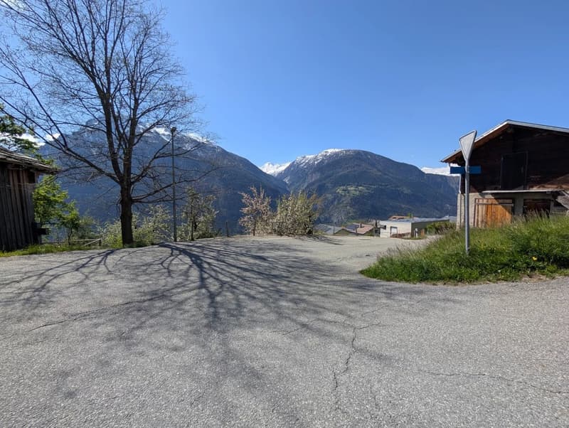 The image shows an outdoor area with a paved parking lot or driveway, surrounded by trees and with a view of snow-capped mountains in the background. The sky is clear and blue, and there are some buildings or structures visible in the distance.