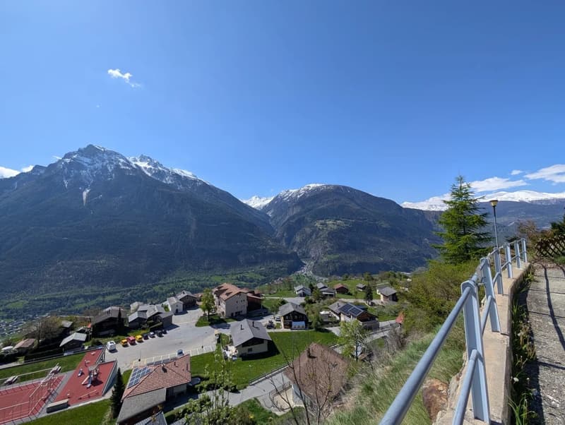 The image shows a picturesque mountain village nestled in a valley surrounded by snow-capped peaks. The village has a mix of traditional and modern buildings, with a winding road and some vehicles visible. The landscape is dominated by the rugged, mountai
