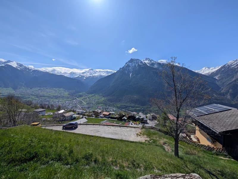 The image shows a scenic mountain landscape with snow-capped peaks, a small village nestled in the valley below, and a clear blue sky with a few clouds. There are solar panels visible on some of the buildings, indicating the use of renewable energy. The f