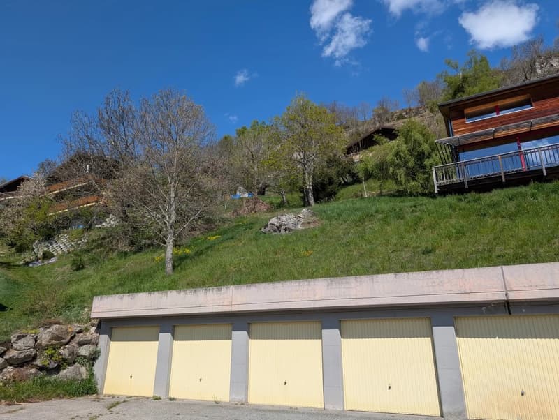 The image shows a building with several garage doors on the ground level, surrounded by a grassy hill with trees and a blue sky with clouds overhead.