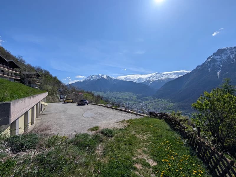 The image shows a scenic mountain landscape with snow-capped peaks in the background. In the foreground, there is a paved parking area surrounded by lush green vegetation and a wooden fence. The sky is a bright blue with some clouds, and the sun is shinin