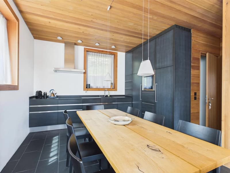 The image shows a modern kitchen and dining area with a wooden ceiling, white walls, and a large wooden dining table. The kitchen features dark gray cabinets and drawers, a stainless steel appliance, and a window. The dining area has a wooden table and ch