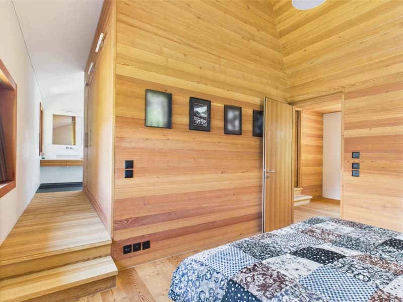 Spacious bedroom with wooden paneled walls and ceiling, framed artwork on the walls, and a patterned bedspread on the bed.