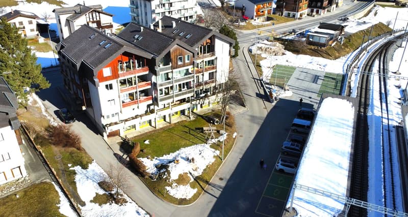 Multi-story apartment building with red and white exterior, balconies, surrounded by snow-covered landscape, with a road and ski lift visible in the background.