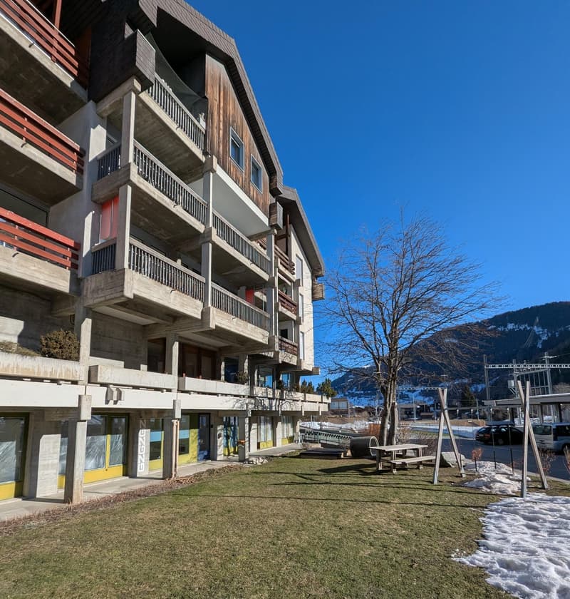 Multi-story building with wooden and concrete exterior, balconies on each floor, ground floor with retail spaces, surrounded by snow-covered grass and trees