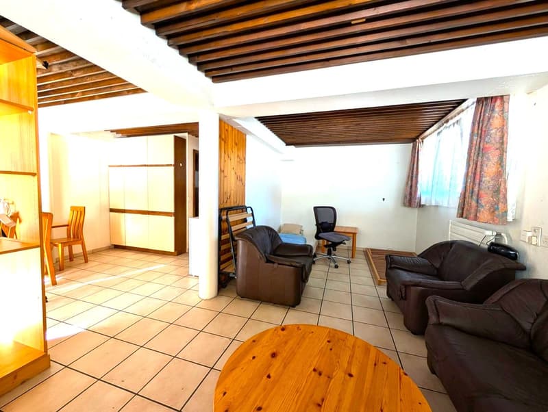 Spacious living room with wooden beamed ceiling, tile flooring, leather sofa and armchair, wooden coffee table, and a desk with a chair. There appears to be a balcony or terrace visible through the window.
