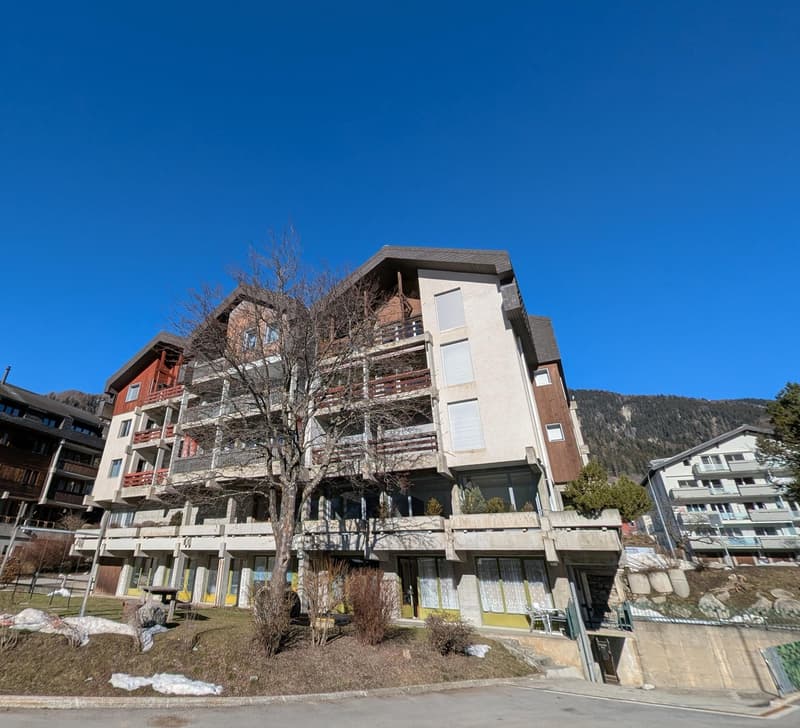Multi-story apartment building with balconies, surrounded by trees and mountains in the background.