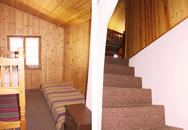 The image shows the interior of a wooden cabin. The walls and ceiling are made of wood panels, creating a cozy and rustic atmosphere. The room has a bed with striped bedding, and there is a staircase leading to another level. The overall impression is of