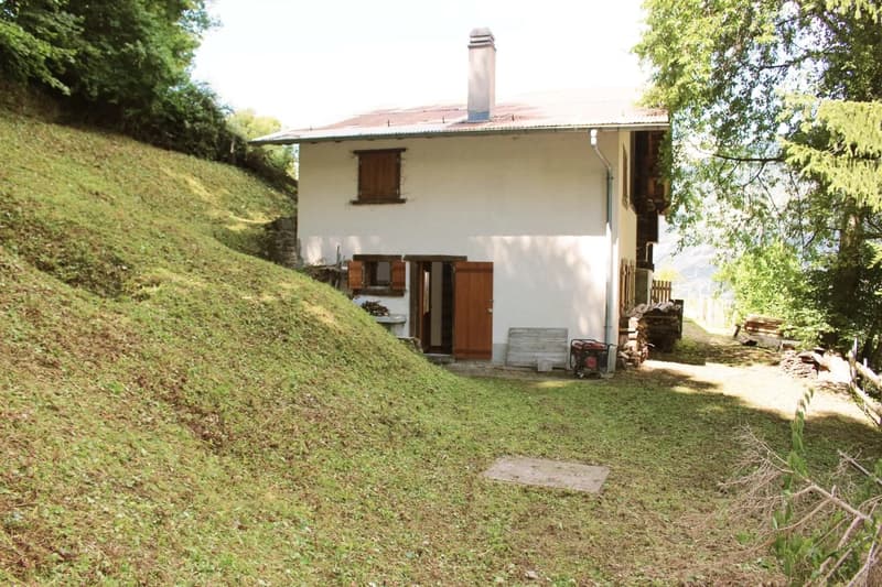 Single-story house with a raised ground floor, surrounded by a grassy hill and trees. The house has wooden windows and a chimney on the roof.