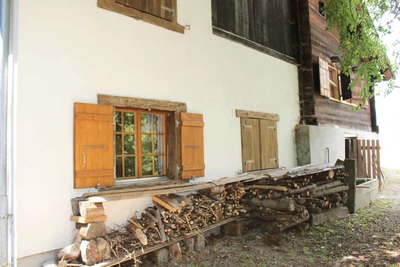 A rustic wooden building with shuttered windows, a stack of firewood on the side, and a natural, wooded environment surrounding it.