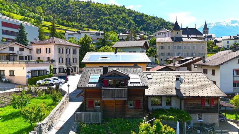 Picturesque town nestled in a lush, green mountainous landscape. The buildings feature a mix of traditional and modern architectural styles, with some having balconies or terraces. There are several multi-story buildings, as well as a church with distinct