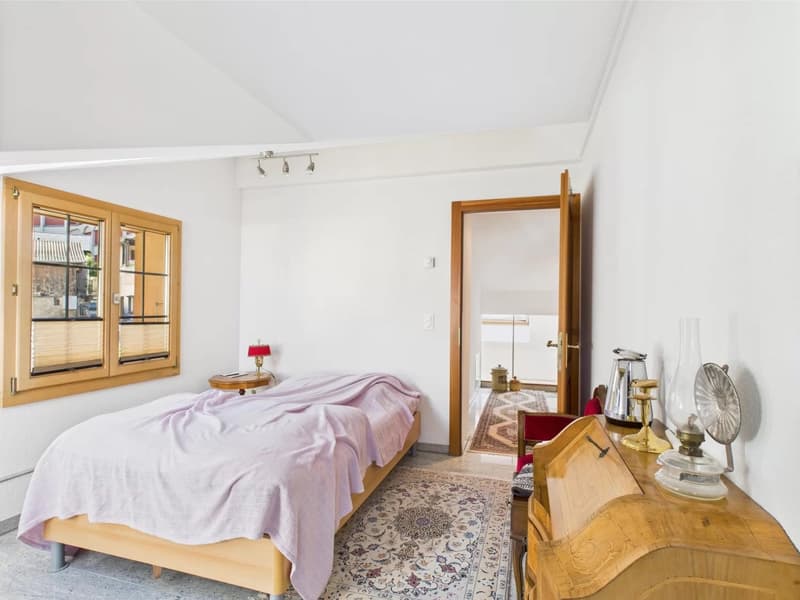 Spacious bedroom with wooden framed windows, wooden furniture including a dresser and bed frame, a patterned area rug, and a cozy lavender-colored bedspread.