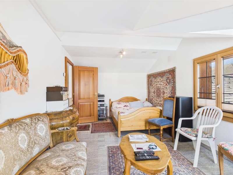 Spacious bedroom with wooden furniture, including a bed, dresser, and armchair. The room also features a patterned rug, curtains, and a balcony or terrace visible through the window.