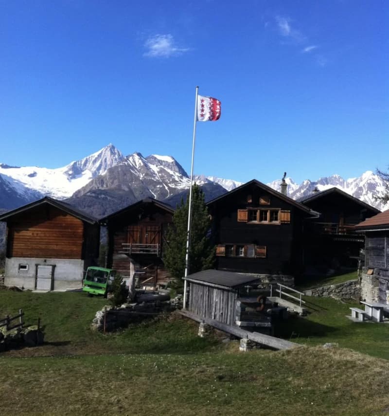 A traditional Swiss mountain chalet with a red and white flag flying, surrounded by snow-capped peaks and a grassy area with a parked vehicle.