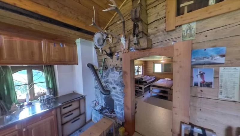 Rustic wooden cabin interior with a fireplace, kitchen area, and sleeping area visible. The cabin has a cozy, outdoorsy feel with mounted antlers and other decor.