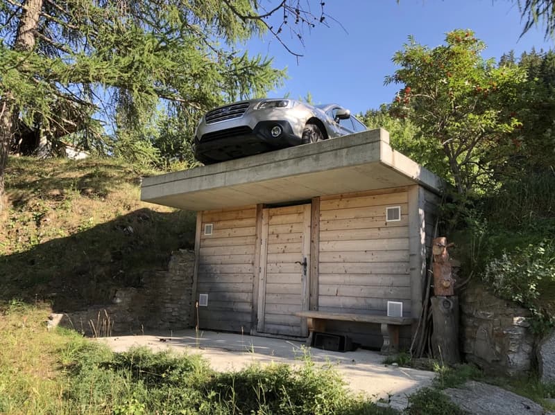 A wooden cabin-like structure with a concrete platform on top, on which a car is parked. The structure is surrounded by lush greenery, including trees and vegetation.