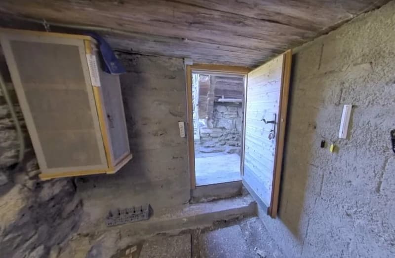 The image shows the interior of an old, dilapidated building. The walls are made of rough, unfinished materials, and the ceiling appears to be made of weathered wooden planks. There is a doorway leading to the outside, which is visible through the open do
