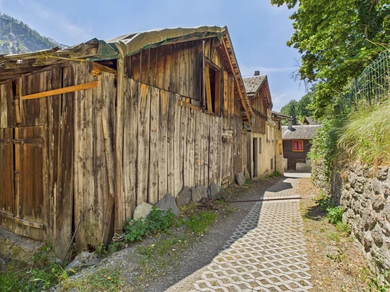 The image shows an old, rustic village with wooden houses and buildings. The houses have weathered, wooden exteriors and appear to be in a rural, mountainous setting with lush greenery surrounding them. There is a paved walkway leading through the village