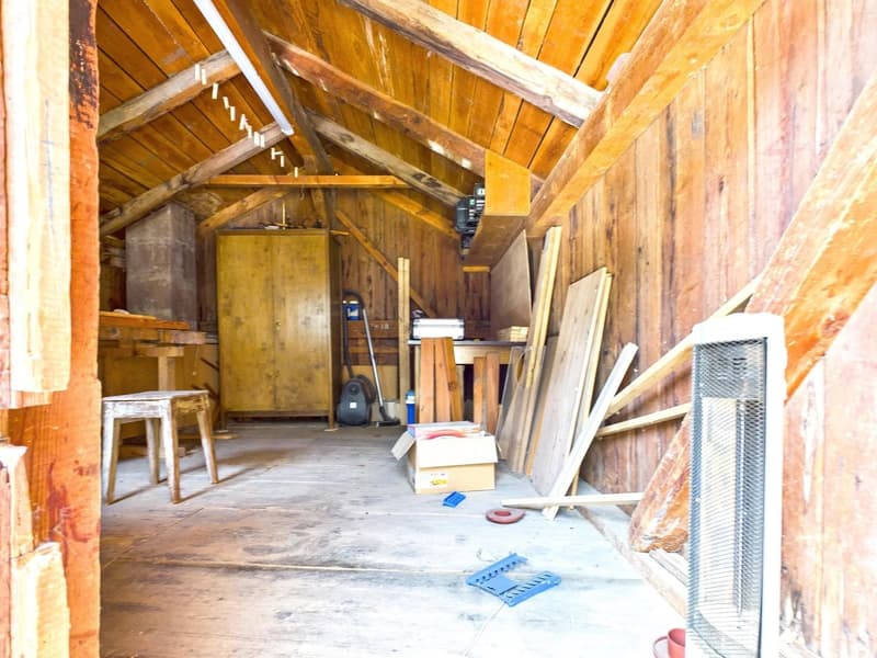 Cluttered storage room with wooden walls and ceiling, various tools and materials scattered on the floor, including a heater, boxes, and wooden planks.