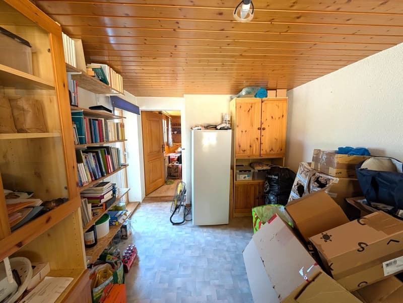 This image shows a cluttered storage room or utility space. The room has a wooden ceiling and walls, with built-in shelves and cabinets along one wall. There is a refrigerator, various boxes and containers, and other miscellaneous items scattered througho