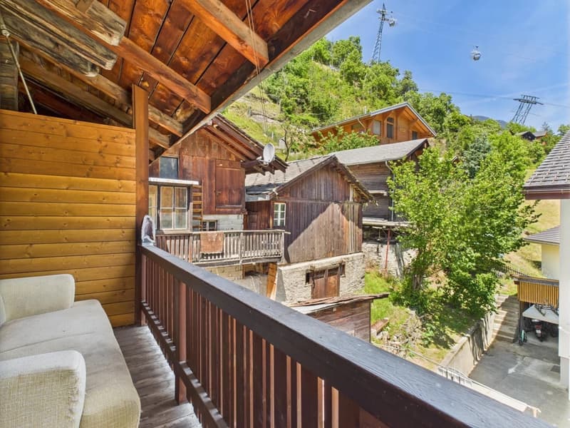 Wooden cabin-style houses with balconies, surrounded by lush greenery and trees. The houses have a rustic, mountain-like appearance.