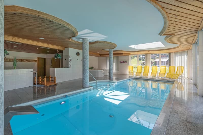 Large indoor swimming pool with wooden ceiling and columns, yellow lounge chairs around the pool, kitchen area visible in the background