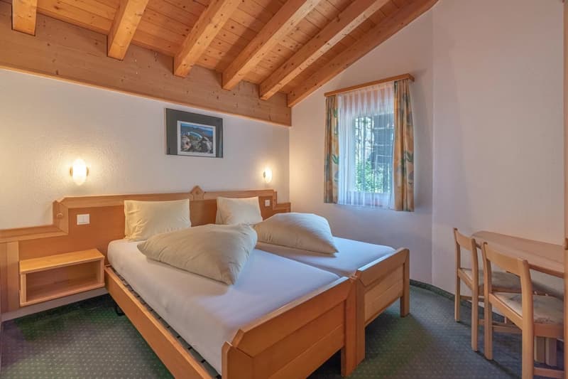 Spacious bedroom with wooden ceiling beams, large windows, wooden furniture including a bed, nightstands, and a chair, and a curtained window providing natural light.