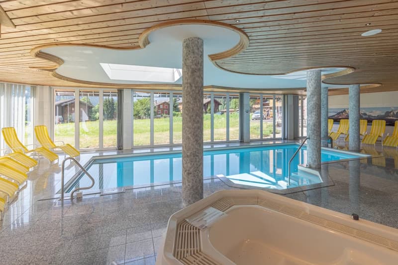 Large indoor swimming pool with yellow lounge chairs, stone columns, wooden ceiling, and large windows overlooking the outdoor scenery.