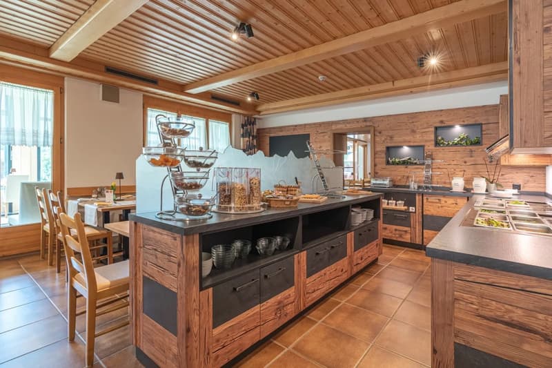 Rustic kitchen with wooden walls and ceiling, large central island with drawers, black countertops, and various kitchen appliances and serving dishes on display