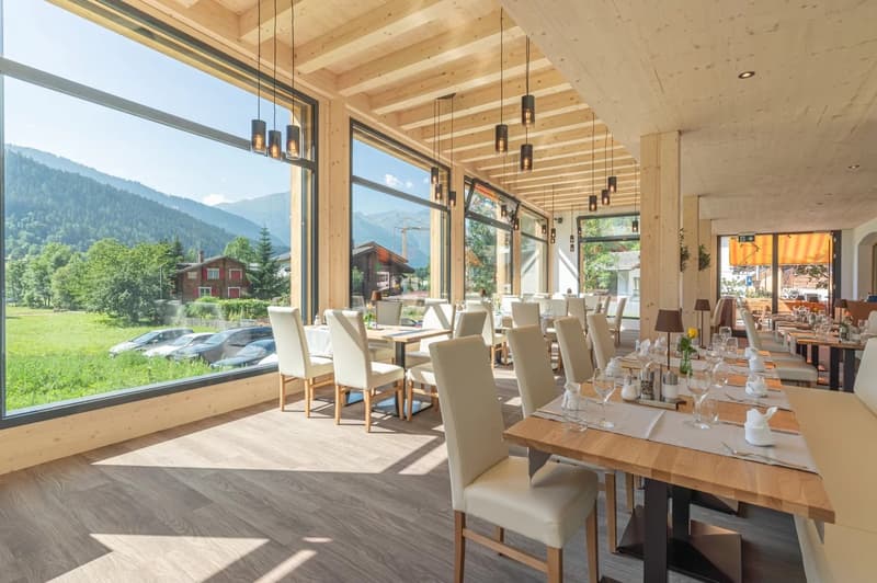 Spacious dining area with large windows offering a scenic mountain view, wooden ceiling beams, pendant lighting fixtures, wooden dining tables and chairs, and a cozy, rustic ambiance.