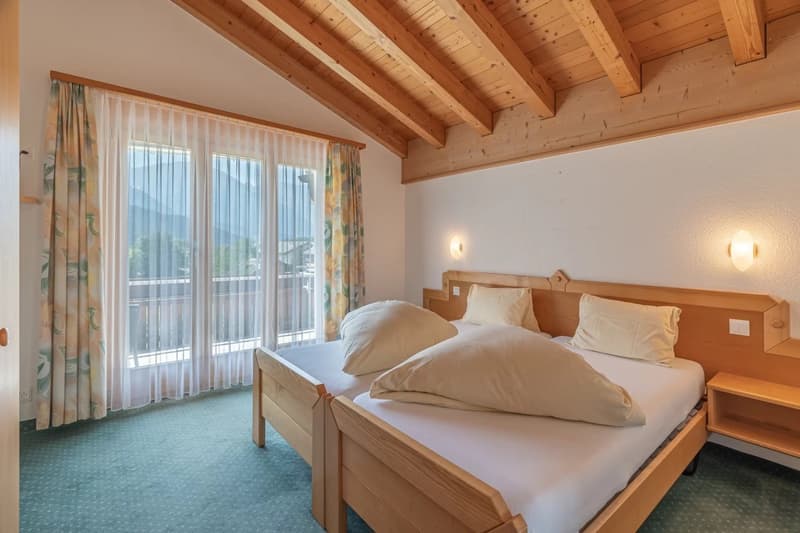 Spacious bedroom with wooden ceiling beams, large windows providing natural light and access to a balcony, wooden furniture including a bed and nightstands, and curtains covering the windows.