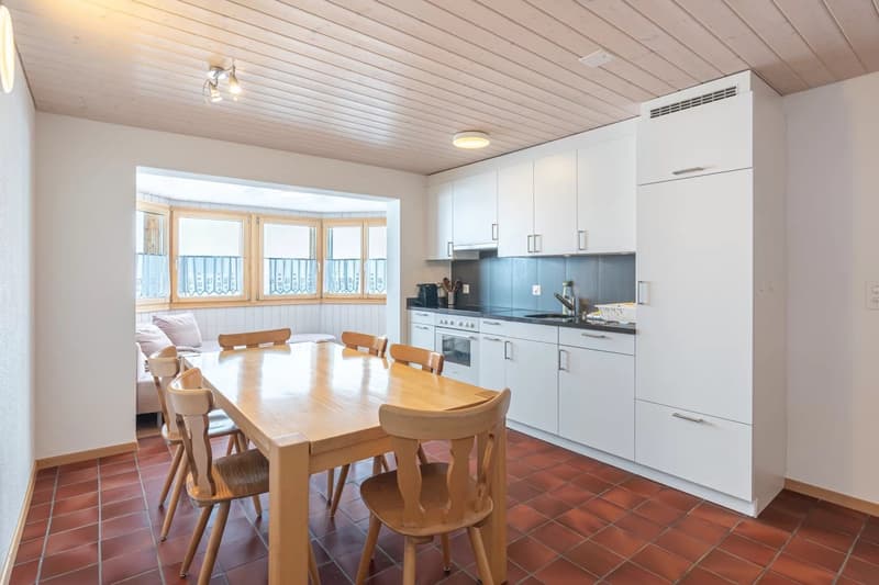 Open-concept kitchen and dining area with large windows, wooden dining table and chairs, white cabinets, and appliances.