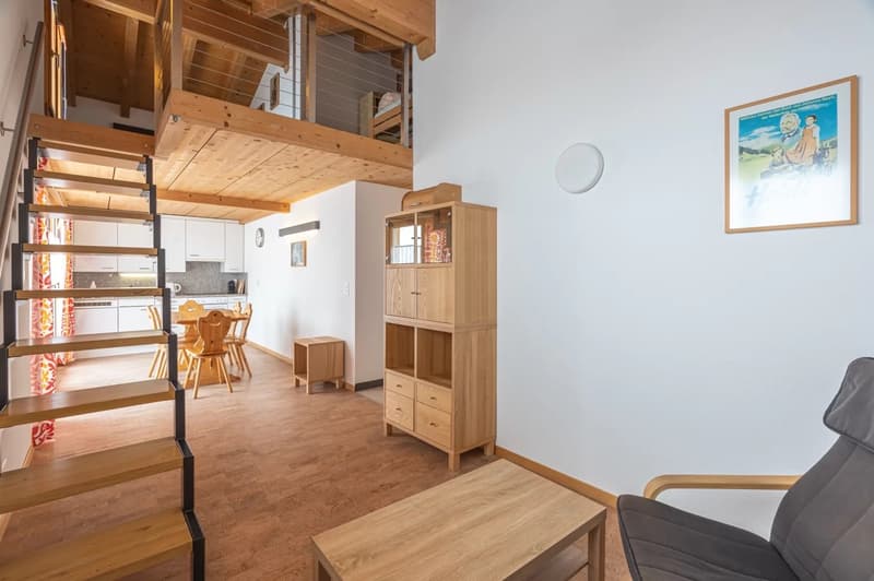 Open-concept studio apartment with wooden beams, loft area, kitchen, dining table, shelving unit, and a framed artwork on the wall.