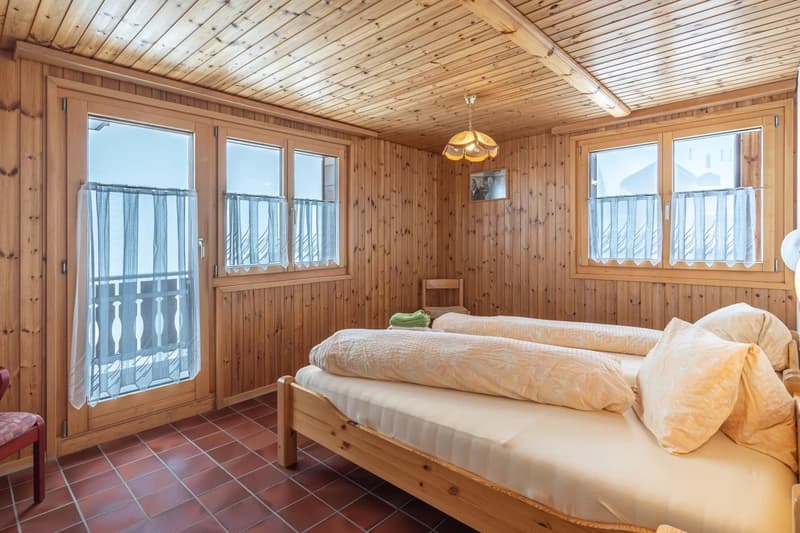 Cozy wooden bedroom with large windows, wooden ceiling, and a balcony. The room features a double bed, wooden furniture, and a decorative light fixture.