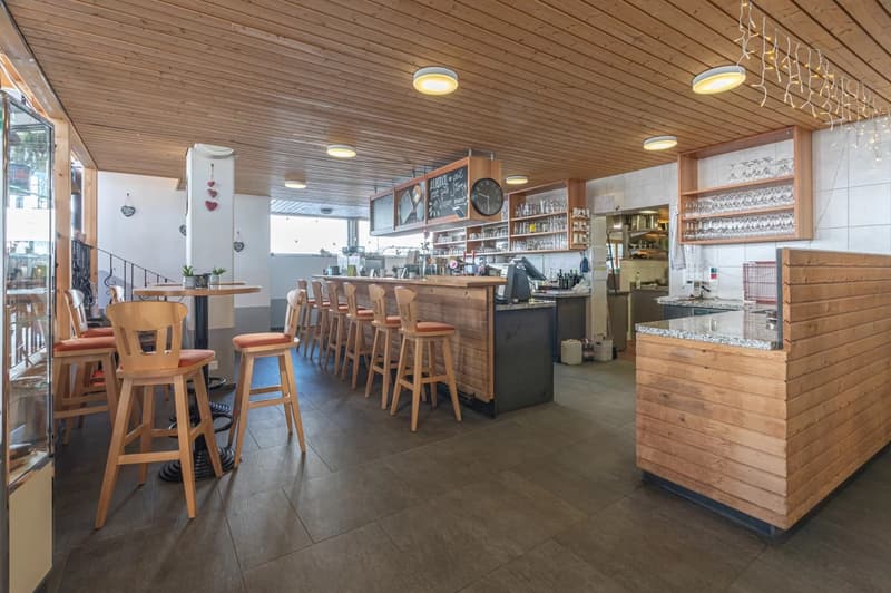 The image shows the interior of a cafe or restaurant. The space has a rustic, wooden decor with a wooden ceiling and walls. There is a long bar counter with stools, and shelves behind the bar displaying various items. The kitchen area is visible in the ba