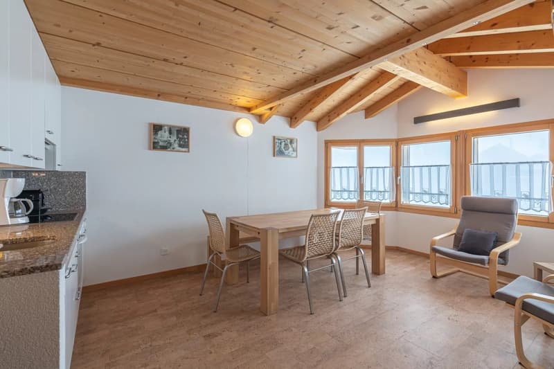 Open-concept living space with wooden ceiling beams, large windows providing natural light, dining table with wicker chairs, and a kitchen area with granite countertops